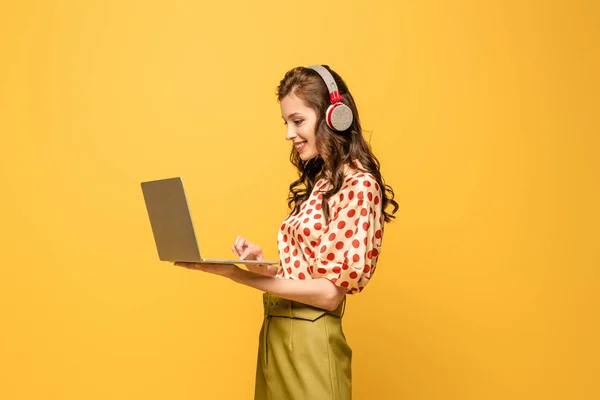Jovem alegre em fones de ouvido sem fio usando laptop isolado no amarelo — Fotografia de Stock