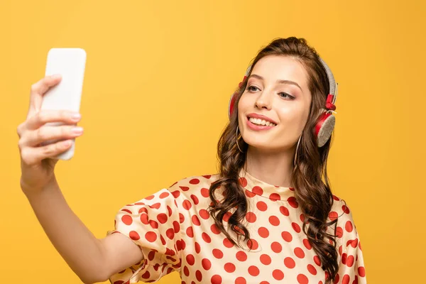 Mujer joven feliz en auriculares inalámbricos tomando selfie en el teléfono inteligente aislado en amarillo - foto de stock