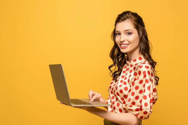 Happy young woman smiling at camera while using laptop isolated on yellow — Stock Photo
