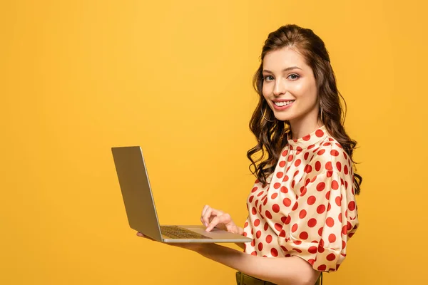 Heureuse jeune femme souriant à la caméra tout en utilisant ordinateur portable isolé sur jaune — Photo de stock