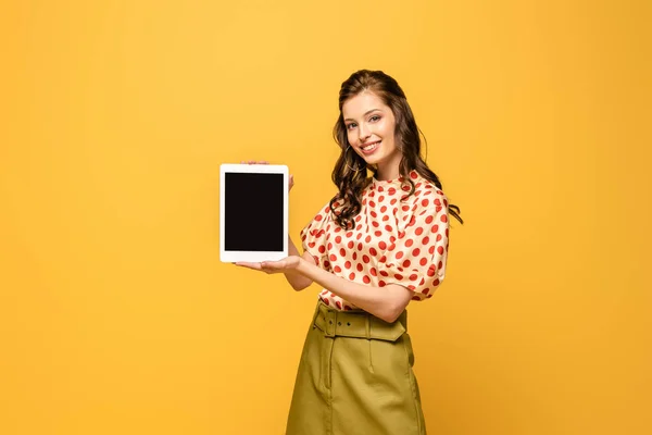 Веселая молодая женщина улыбается в камеру, показывая цифровой планшет с чистым экраном изолированы на желтый — стоковое фото