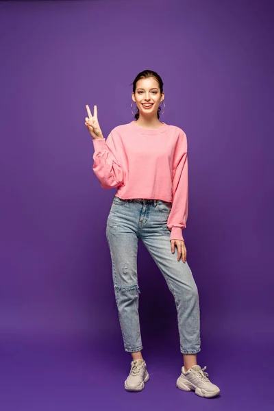 Visão de comprimento total da menina atraente sorrindo para a câmera enquanto mostra gesto de vitória no fundo roxo — Fotografia de Stock