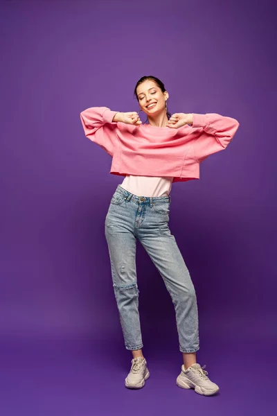 Full length view of happy girl stretching with closed eyes while standing on purple background — Stock Photo
