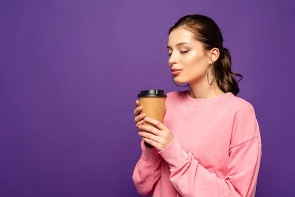 Sognante, ragazza positiva che tiene il caffè per andare mentre in piedi con gli occhi chiusi isolati sul viola — Foto stock