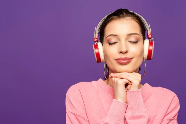Chica sonriente y soñadora escuchando música en auriculares inalámbricos con los ojos cerrados aislados en púrpura - foto de stock