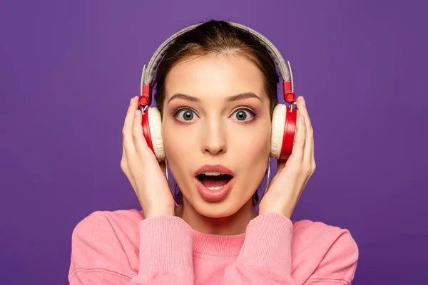 Shocked girl looking at camera while touching wireless headphones isolated on purple — Stock Photo