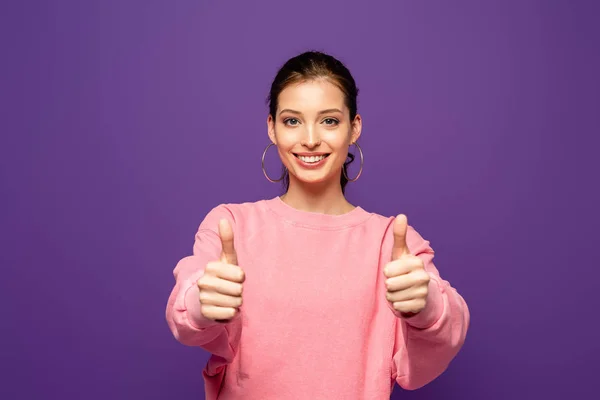Menina atraente sorrindo para a câmera enquanto mostra os polegares isolados no roxo — Fotografia de Stock