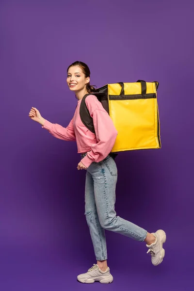 Bastante mensajero corriendo mientras lleva caja termo y sonriendo a la cámara sobre fondo púrpura - foto de stock