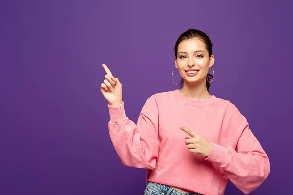 Cheerful girl smiling at camera while pointing with fingers isolated on purple — Stock Photo