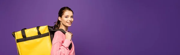 Panoramic shot of pretty courier carrying thermo box and smiling at camera isolated on purple — Stock Photo