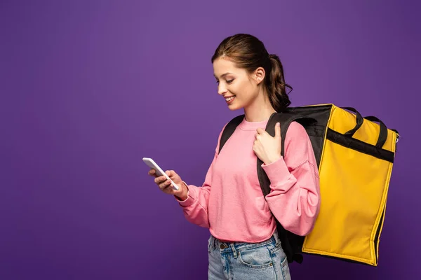 Pretty, smiling courier carrying thermo box and using smartphone isolated on purple — Stock Photo