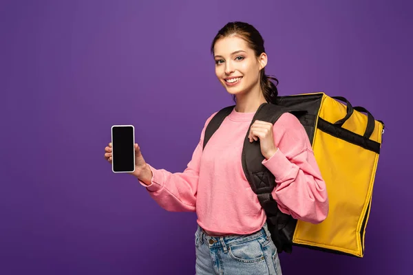 Atractivo mensajero que lleva caja termo y la celebración de teléfono inteligente con pantalla en blanco aislado en púrpura - foto de stock