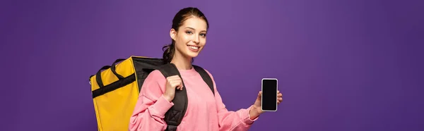 Plano panorámico de mensajero bonito que lleva la caja termo y la celebración de teléfono inteligente con pantalla en blanco aislado en púrpura - foto de stock