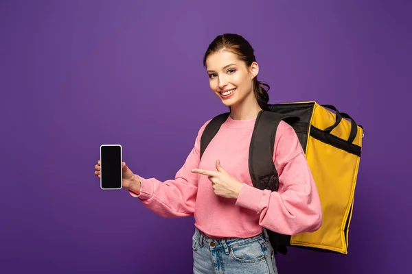Bonito mensajero llevando caja termo y apuntando con el dedo al teléfono inteligente con pantalla en blanco aislado en púrpura - foto de stock