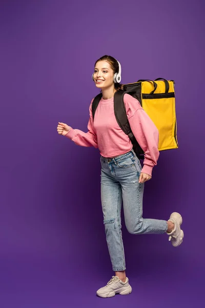 Coursier joyeux fonctionnant dans des écouteurs tout en portant thermo boîte sur fond violet — Photo de stock