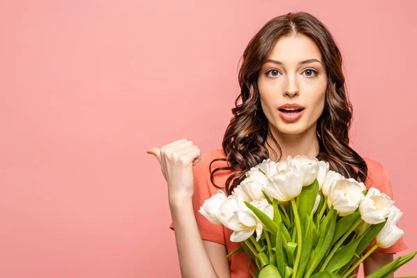 Sorprendida joven mujer mirando a la cámara y apuntando con el pulgar mientras sostiene tulipanes blancos aislados en rosa - foto de stock