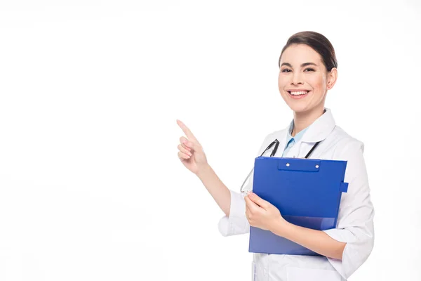 Médico sonriente con estetoscopio y portapapeles sonriendo a la cámara mientras señala aislado en blanco - foto de stock