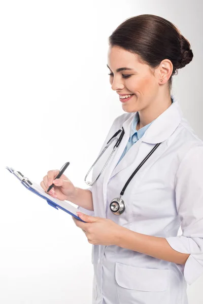 Vista lateral del médico sonriente escribiendo en portapapeles aislado en blanco - foto de stock