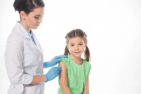 Pediatra femenina con algodón frotando hombro de niño sonriente aislado en blanco — Stock Photo