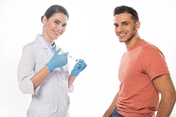 Vista lateral del médico sosteniendo la jeringa y la vacuna y sonriendo a la cámara cerca del paciente aislado en blanco - foto de stock