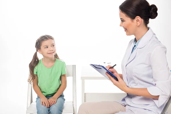 Pediatra sonriente con portapapeles mirando a un niño en silla aislado en blanco — Stock Photo