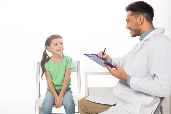 Smiling pediatrician looking at kid and writing on clipboard isolated on white — Stock Photo