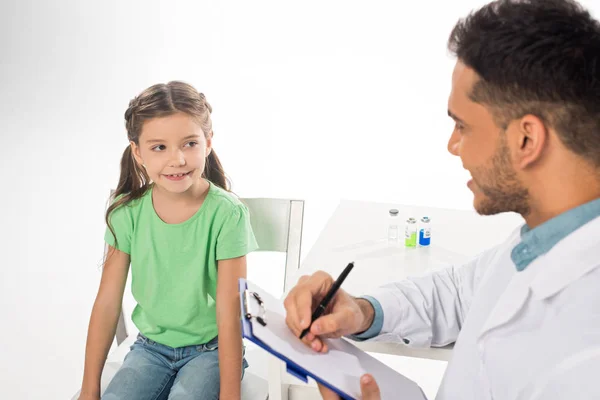 Selektiver Fokus des lächelnden Kindes beim Anblick des Kinderarztes mit Klemmbrett isoliert auf Weiß — Stockfoto