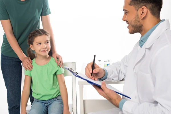 Smiling pediatrician with clipboard looking at kid near mother isolated on white — Stock Photo