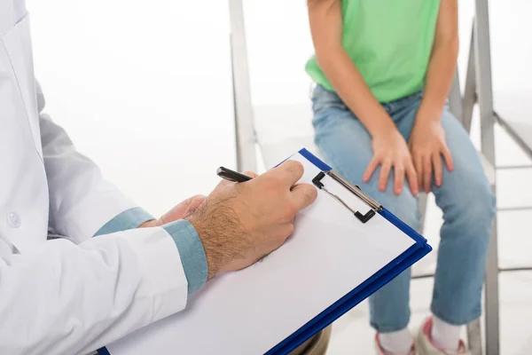 Selective focus of pediatrician writing on clipboard near kid on chair on white background — Stock Photo