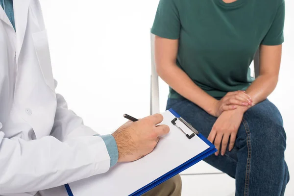 Cropped view of doctor writing on clipboard near patient on chair isolated on white — Stock Photo