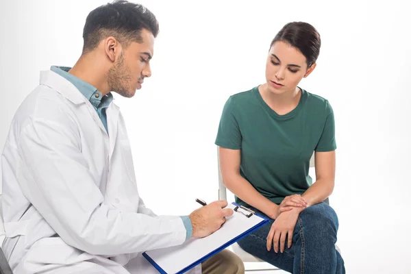 Guapo doctor escribiendo en portapapeles cerca de paciente femenina aislado en blanco - foto de stock