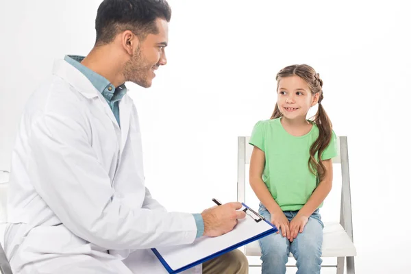 Smiling pediatrician with clipboard looking at kid on chair isolated on white — Stock Photo