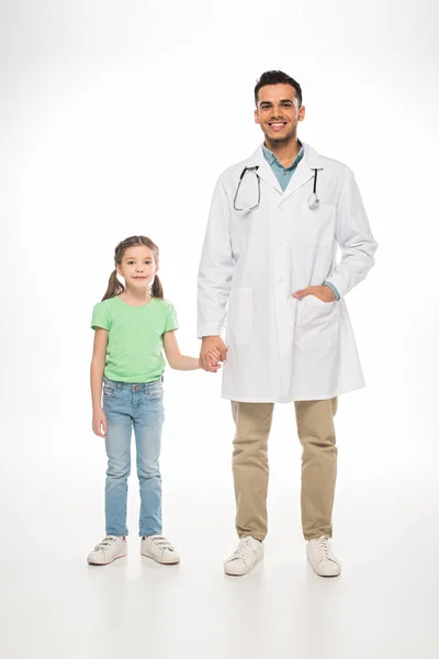 Pleine longueur de pédiatre souriant et enfant tenant la main et souriant à la caméra sur fond blanc — Photo de stock