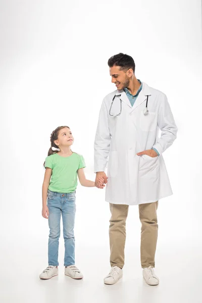 Полная длина ребенка и педиатра улыбаются друг другу и держатся за руки на белом фоне — стоковое фото