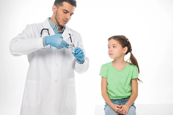 Handsome pediatrician picking up vaccine in syringe near kid on table isolated on white — Stock Photo