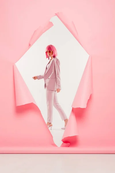 Modèle élégant en costume tendance, lunettes de soleil et perruque rose posant en papier déchiré, sur blanc — Photo de stock
