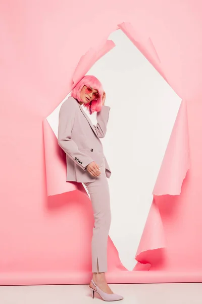Modèle à la mode en costume, lunettes de soleil et perruque rose posant en papier déchiré, sur blanc — Photo de stock