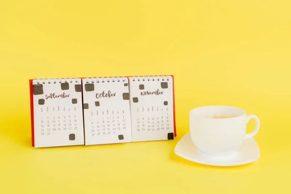 Calendario con meses de otoño y taza de café sobre fondo amarillo - foto de stock