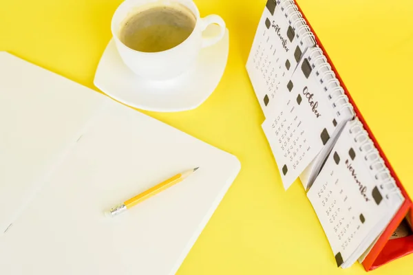 Blick auf leeres Notizbuch in der Nähe von Kaffeetasse und Kalender auf gelbem Hintergrund — Stockfoto