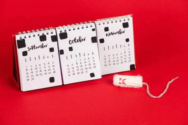 Calendário e tampão higiênico com expressão facial triste no fundo vermelho — Fotografia de Stock