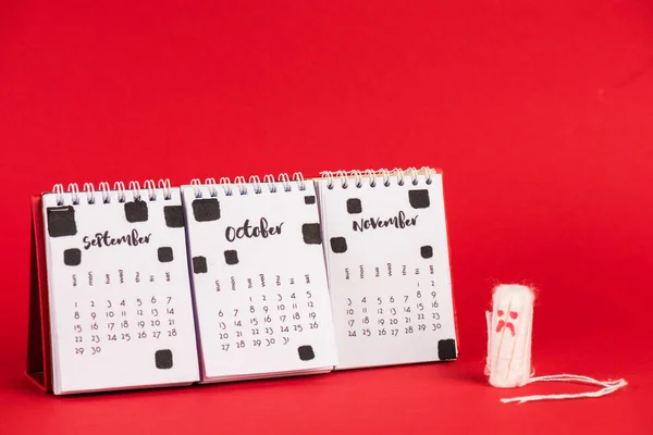 Tampão higiênico com expressão facial triste perto do calendário com meses de outono no fundo vermelho — Fotografia de Stock