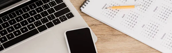 Panoramic shot of smartphone on laptop with calendar and pencil on wooden background — Stock Photo