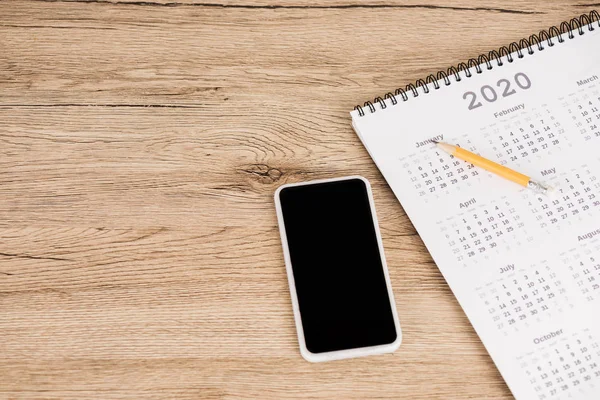 Vista de alto ángulo del teléfono inteligente con pantalla en blanco y calendario de 2020 año con lápiz sobre fondo de madera - foto de stock