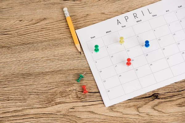 Visão de alto ângulo de lápis, alfinetes de escritório e calendário de abril no fundo de madeira — Fotografia de Stock