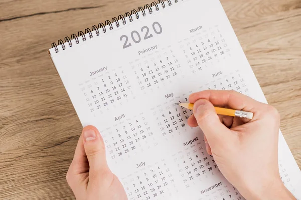 Vista recortada del hombre que observa la fecha con lápiz en el calendario sobre fondo de madera - foto de stock