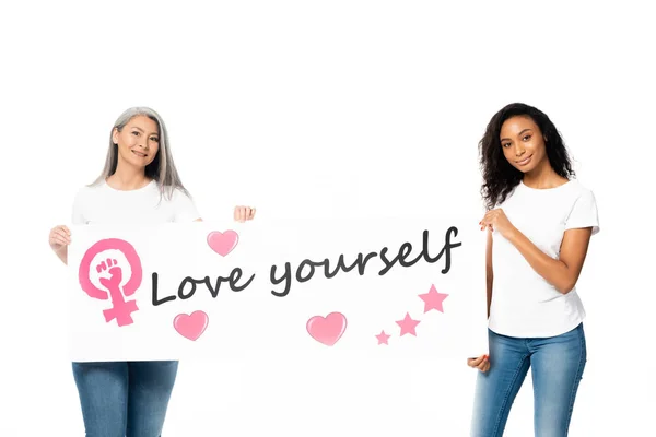 Happy african american and asian women holding placard with love yourself lettering and women power sign isolated on white — Stock Photo