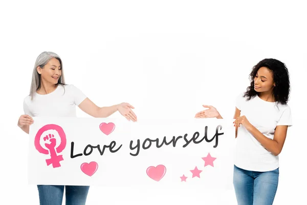 Щасливі афро-американські та азіатські жінки, що вказують руками на плакаті з любов'ю, саджають ізольовані на білому — стокове фото