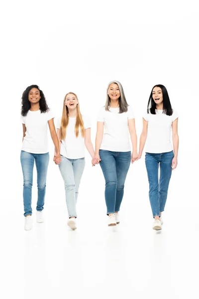 Heureuses femmes multiculturelles en jeans denim marchant et se tenant les mains isolées sur blanc — Photo de stock