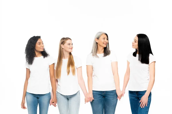 Mujeres multiculturales felices en vaqueros tomados de las manos aislados en blanco - foto de stock