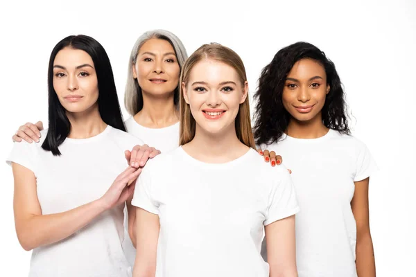 Glückliche multikulturelle Frauen in weißen T-Shirts, die isoliert auf weiß in die Kamera schauen — Stockfoto